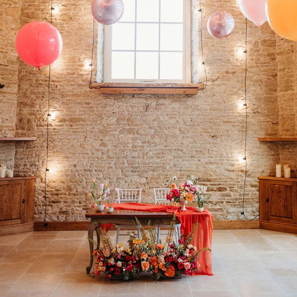 Merriscourt wedding venue reception room with metallic pink, white, orange and yellow balloons, wedding fairy lights decorations and wooden table with orange table runner and mixed seasonal flower arrangements 