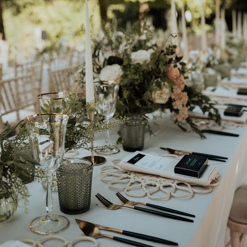 Back and gold cutlery, black and white menu card and white wicker place mats with green glassware, gold rimmed wine glasses and neutral flowers
