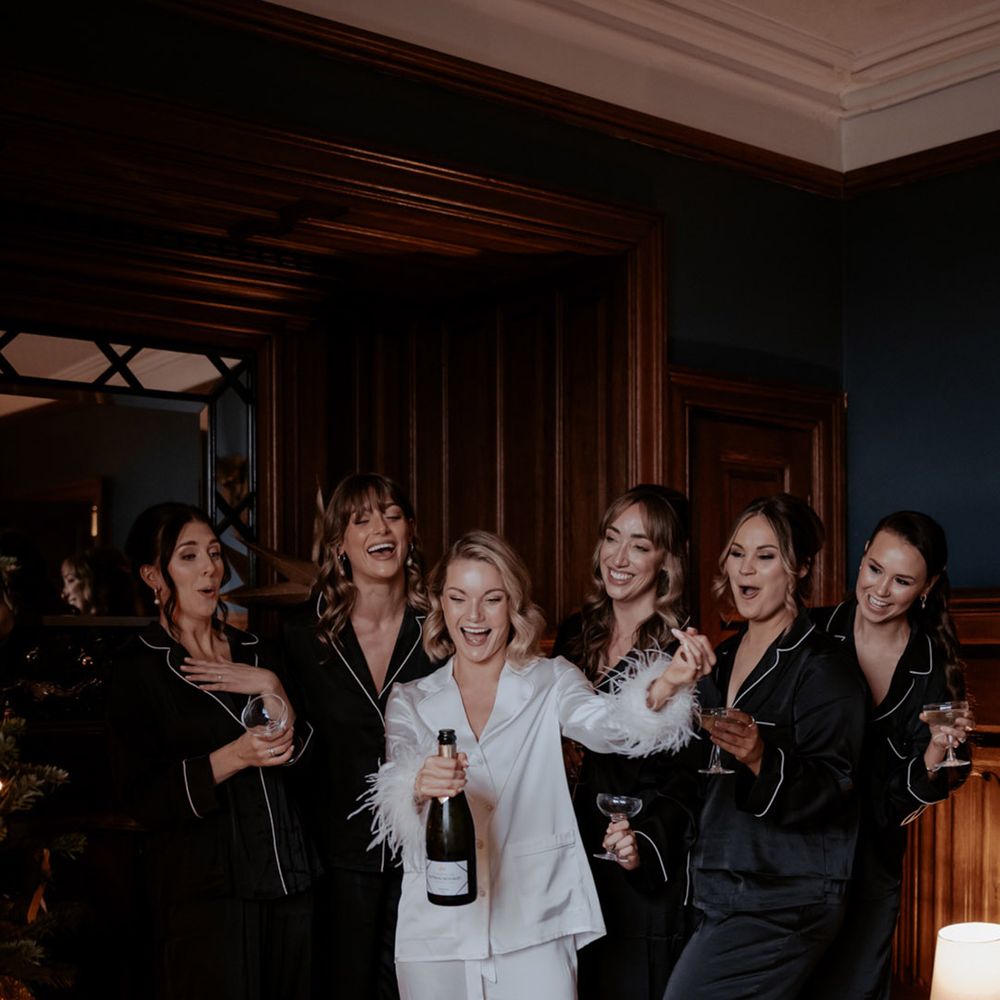 Ladies in black satin pyjamas with bride in white feather pyjamas popping bottle of champagne