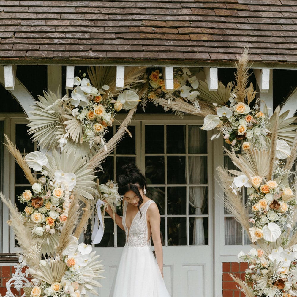 Bride at Berwick lodge wearing a plunge neck wedding gown with tulle skirt, under an arch of dried palms, pampas grass, white anthuriums, roses and orchids