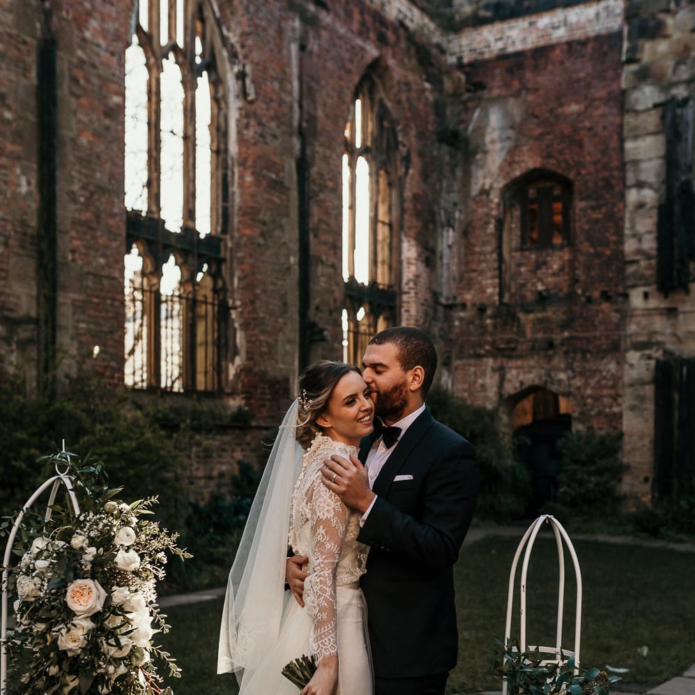 Groom in a black tuxedo kissing his brides head in a lace and tulle fitted wedding dress at St Luke's Bombed Out Church in Liverpool 