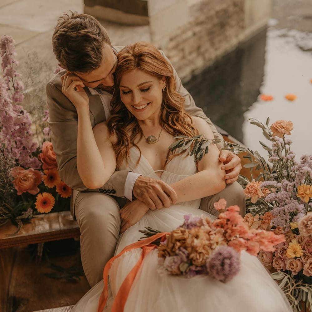 Bride with long wavy red hair sitting on a boat with her groom surrounded by pastel orange, pink and lilac wedding flowers at Euridge Manor boat house 