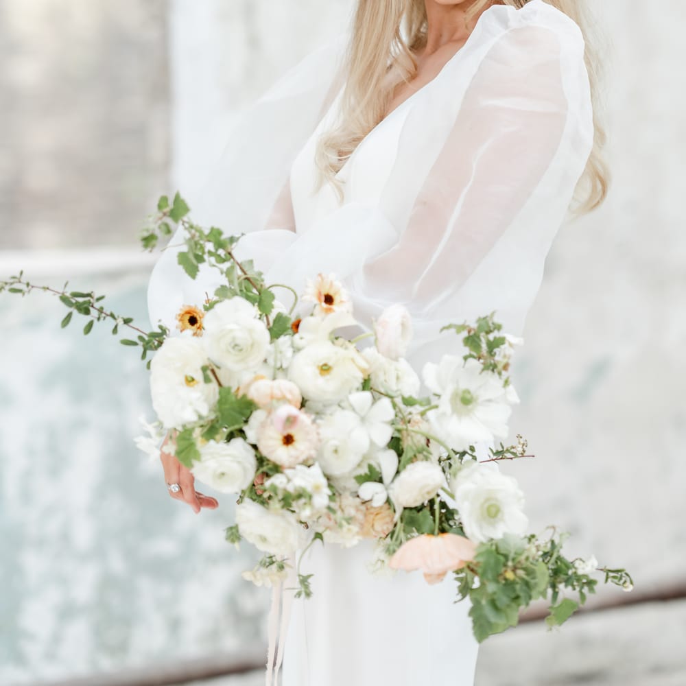 Bride in a sheer long sleeve wedding dress with straight hair, a halo headdress and red lipstick holding a romantic white wedding bouquet 