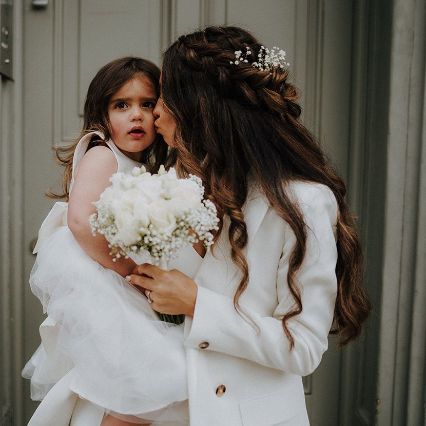 Bride in a trouser suit holding her daughter and a white rose bouquet 