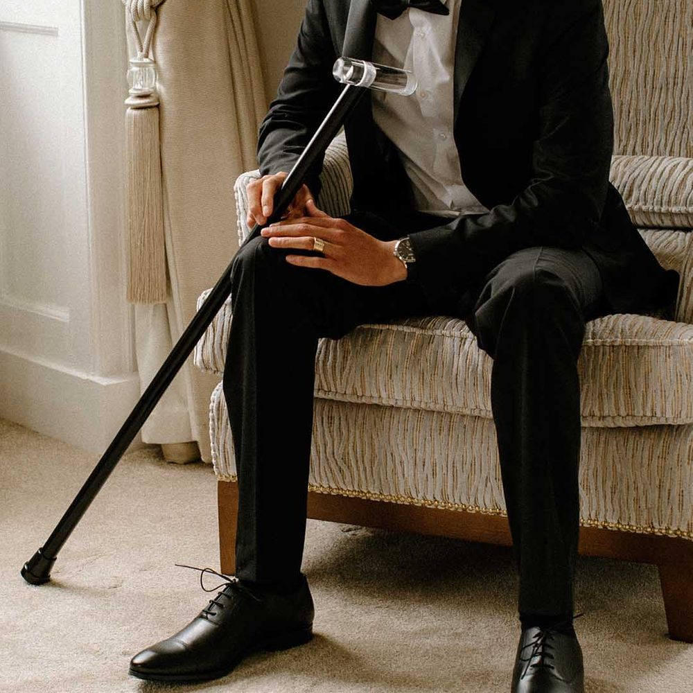 Interable groom in a black tuxedo sitting on a chair at Bourton Hall with his walking stick 