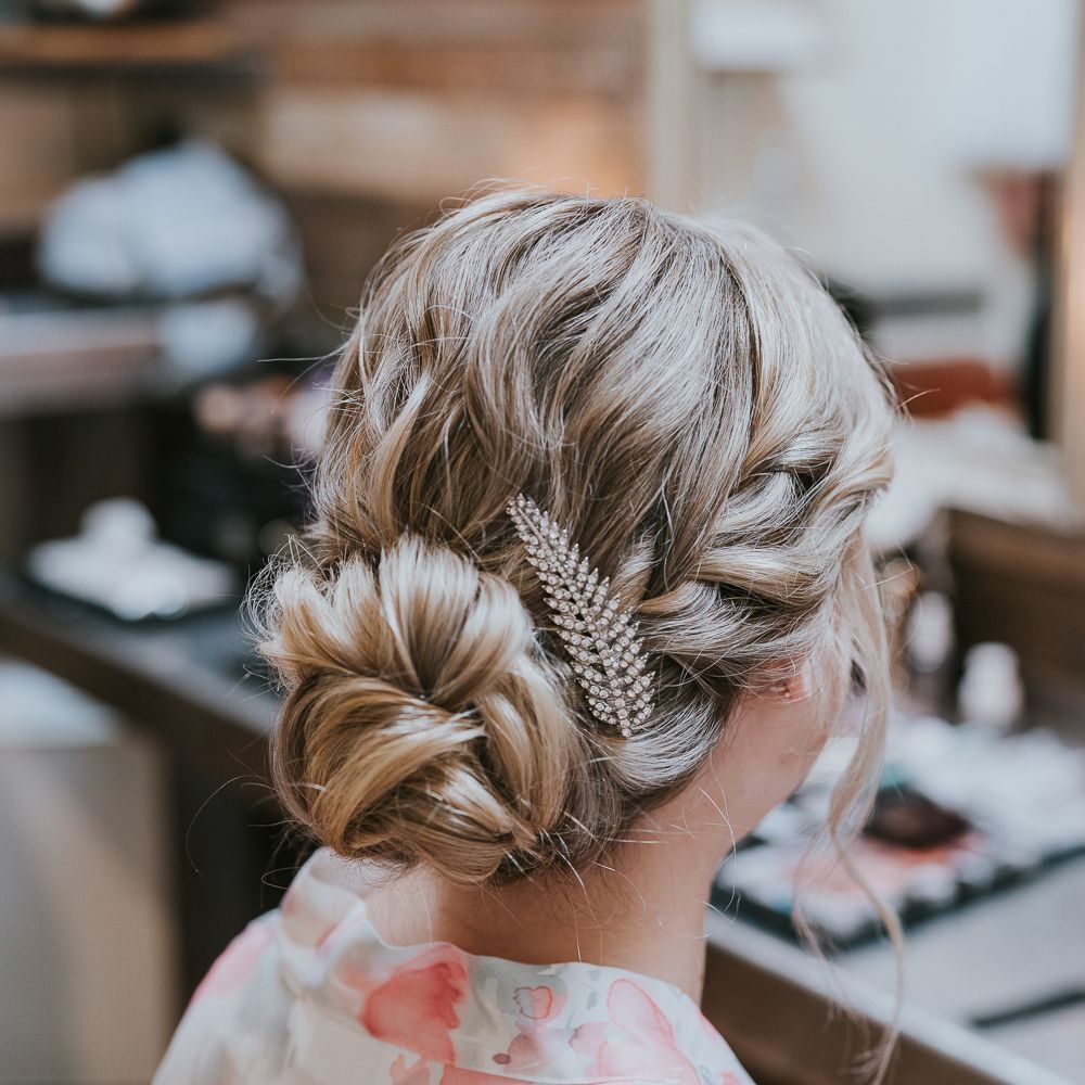 Bride with plaited bun and leaf hair clip sits at mirrors before Tythe Barn wedding with barn wedding flowers