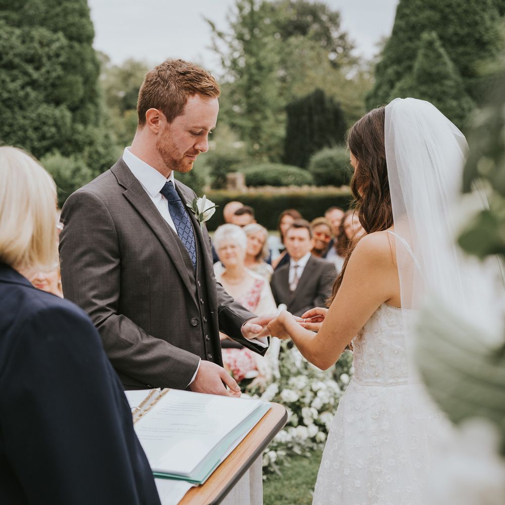 Bride in white Pronovias wedding dress and veil puts ring on hand of groom in grey three piece suit, blue tie and white rose buttonhole during outdoor wedding ceremony