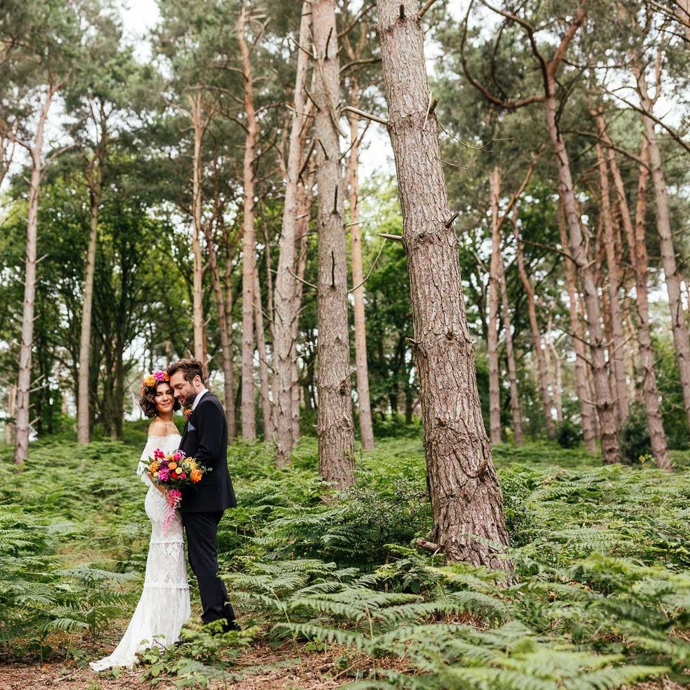 Bride in off the shoulder Grace Loves Lace wedding dress and flower crown holding colourful bridal bouquet stands with groom in black suit in woodland after outdoor wedding ceremony at The Bridal Barn in Claverley