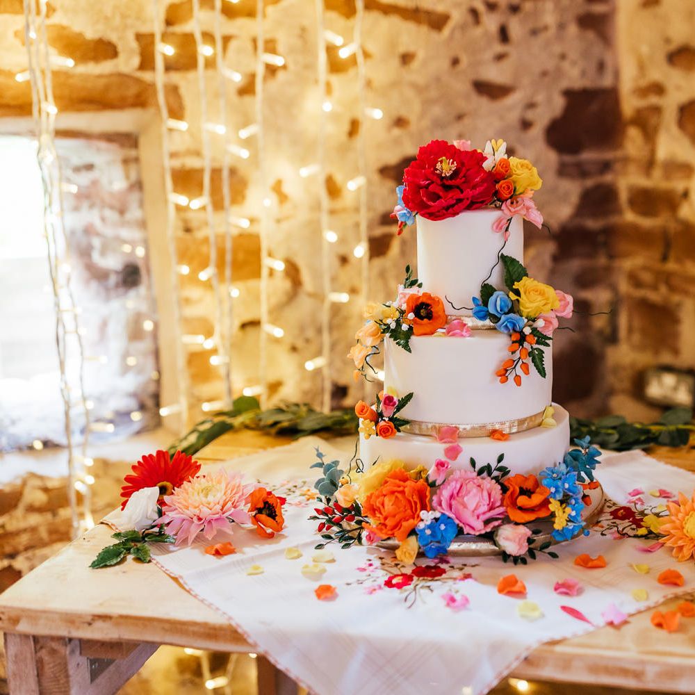 White three tier wedding cake with multicoloured floral decorations on wooden table at The Bridal Barn wedding reception