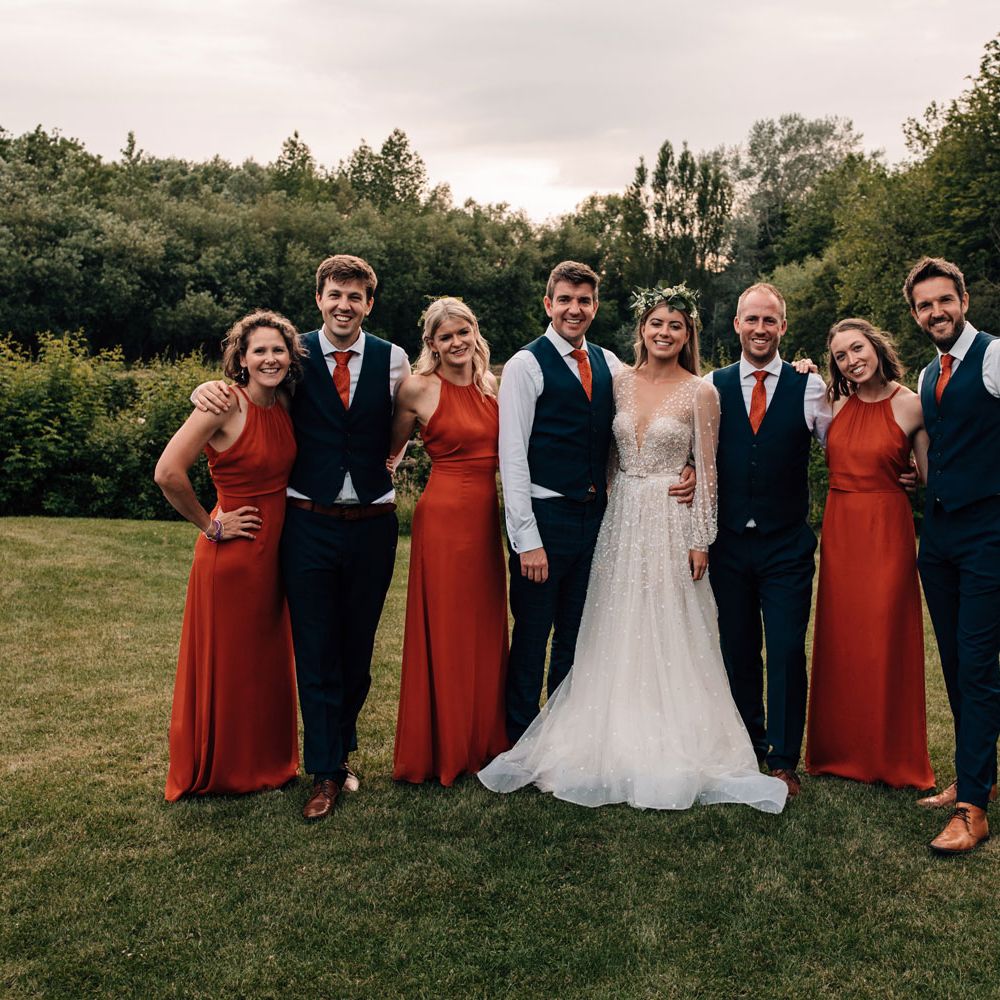 Bride in long mesh sleeve Julie Vino wedding dress stands with bridesmaids in red bridesmaid dresses and groomsmen in dark waistcoats and red ties at tipi wedding reception