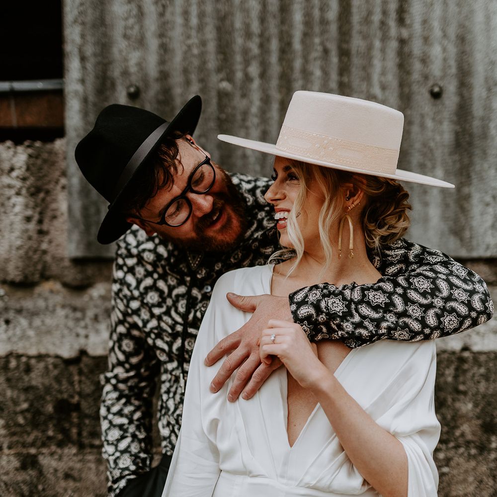 Groom wraps his arm around his bride who wears bridal hat and jumpsuit with cape on their wedding day