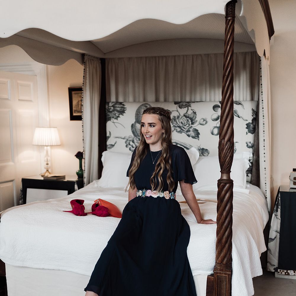 Bridesmaid in navy bridesmaid dress with floral embroidered and blue heels sits on bed before wedding