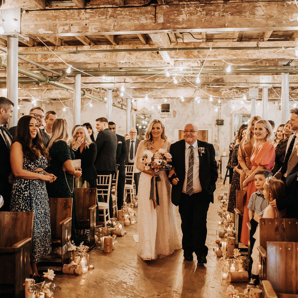 Bride in white pearl wedding dress with sleeves and pastel mixed bouquet with roses walks down the aisle with man in dark suit and striped tie at industrial style wedding