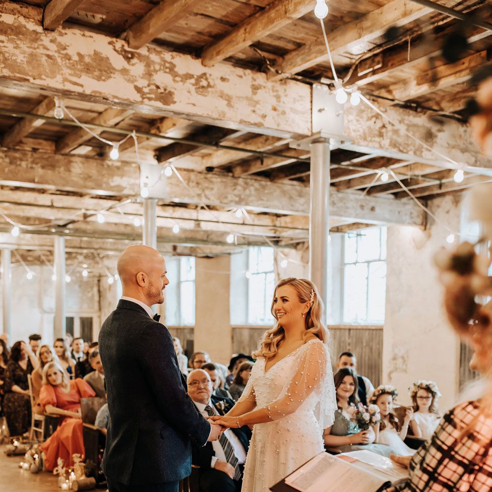 Bride in white v neck wedding dress with pearls stands with groom in navy suit during industrial style wedding ceremony
