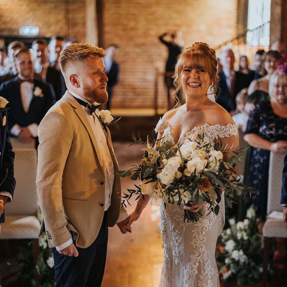 Groom in beige suit jacket with bow tie stands with bride at the end of the altar with bride in Maggie Sottero lace wedding dress