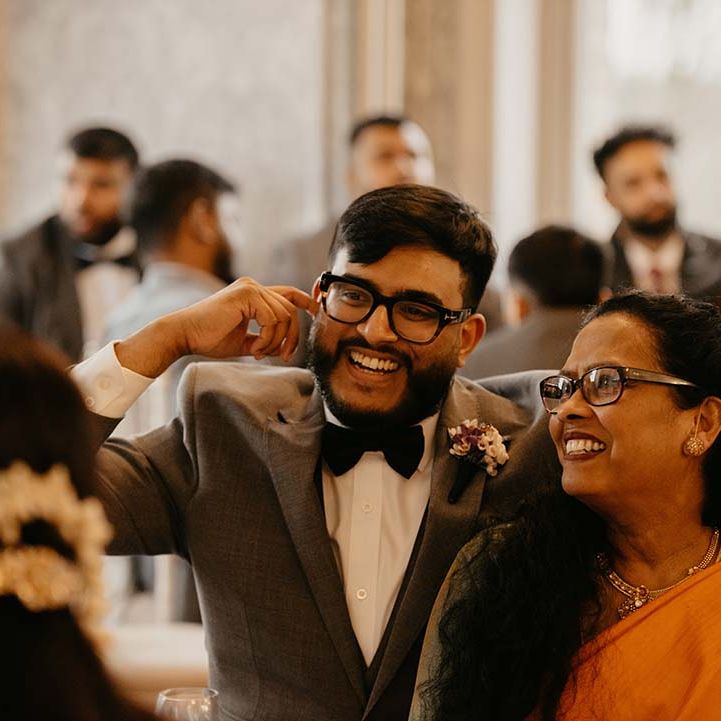 Groom in dark grey suit and black bow tie embracing wedding guest in vibrant orange saree 