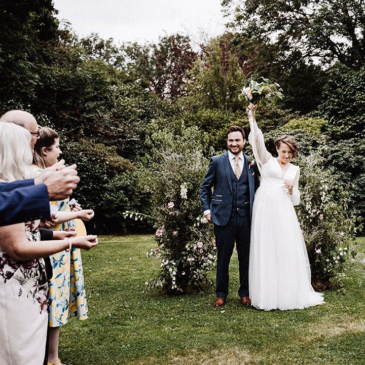 Bride in long sleeve wedding dress raises her bouquet in celebration with groom in blue suit at the altar