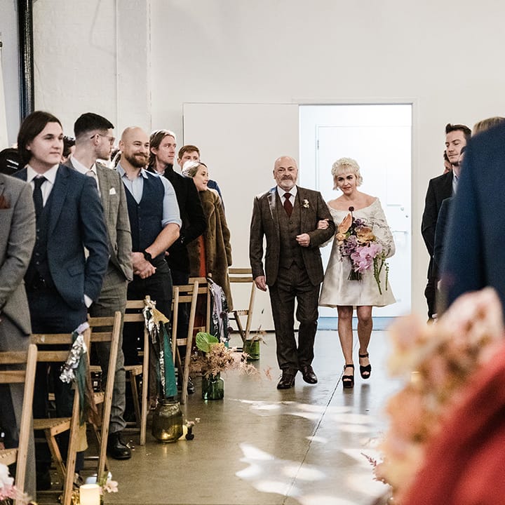 Father of the bride in checkered three piece suit walks the bride in a short wedding dress down the aisle