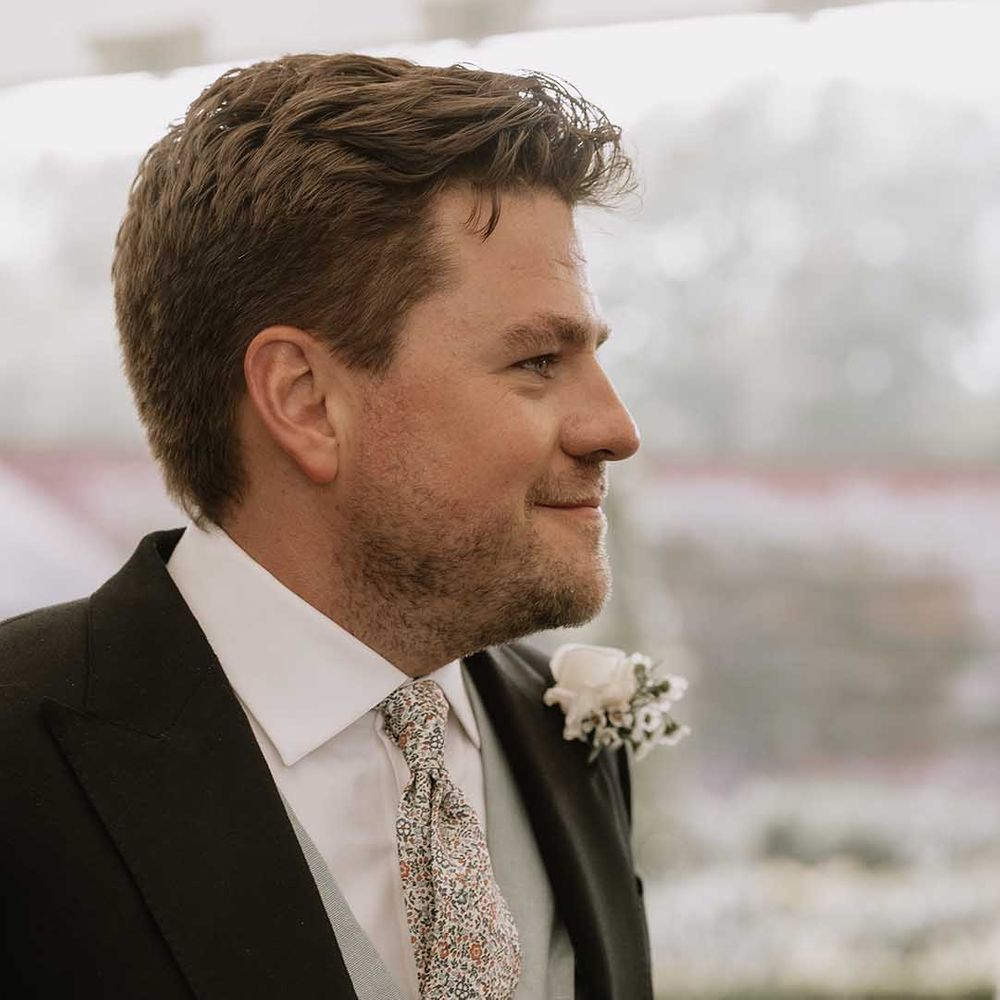 Groom in a green waistcoat and floral patterned tie and white flower buttonhole smiles as he sees the bride for the first time 