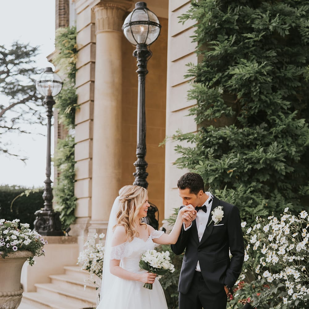 Groom in black tie kisses the bride's hands as she wear an off the shoulder wedding dress and white flower bouquet 
