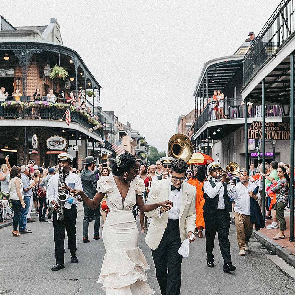 Black bride in a fitted wedding dress with ruffle marching down the street with her groom in a white tuxedo jacket followed by a brass band