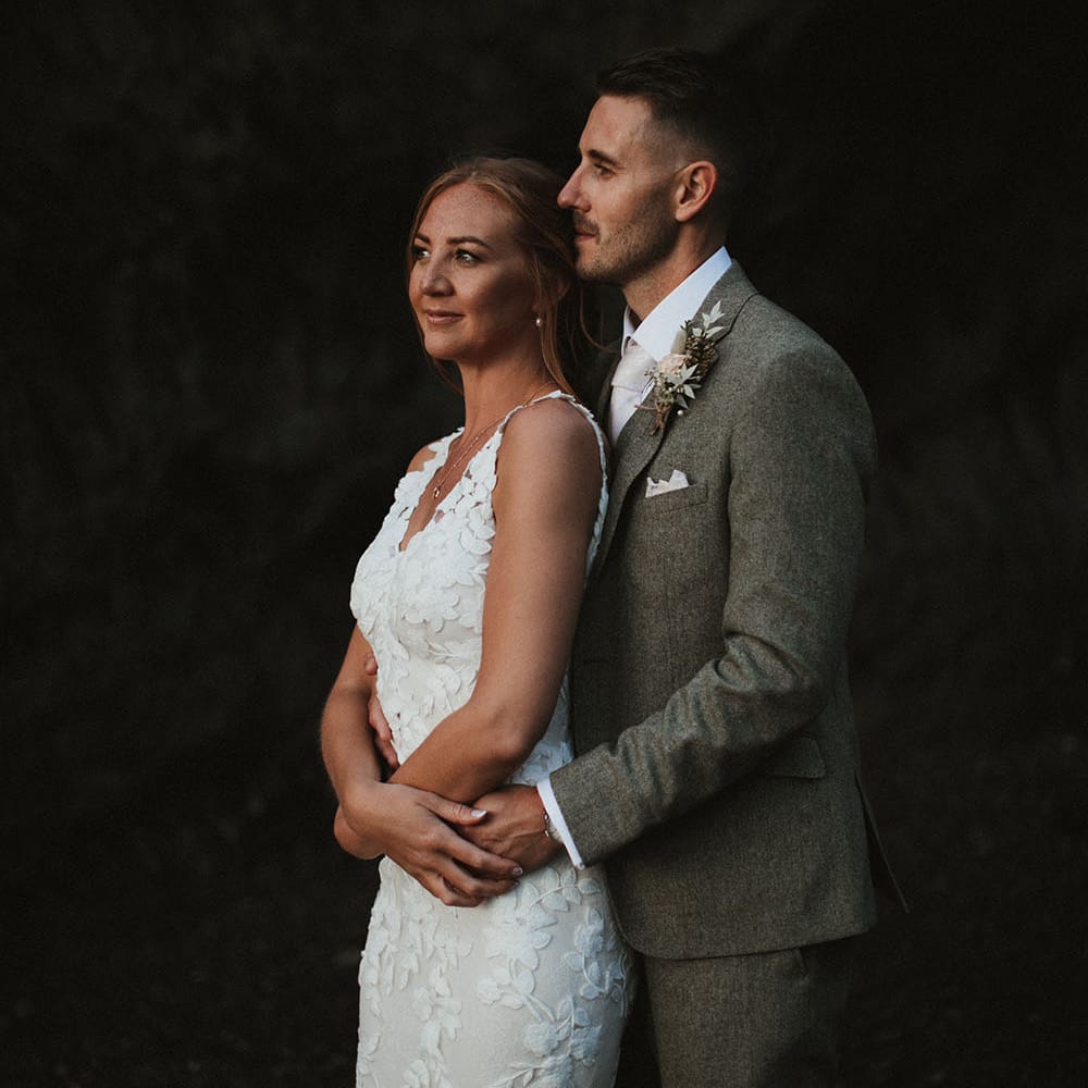 Bride wears fitted floral embellished Enzoani wedding dress as her groom stands behind her wearing green suit with white pocket square and floral buttonhole