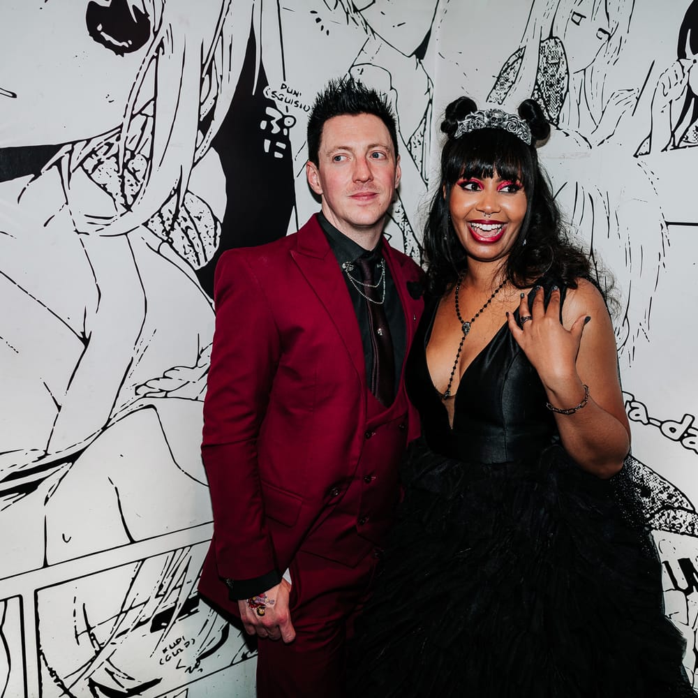 Bride wearing black ruffled wedding dress stands with her groom in front of anime styled wall 