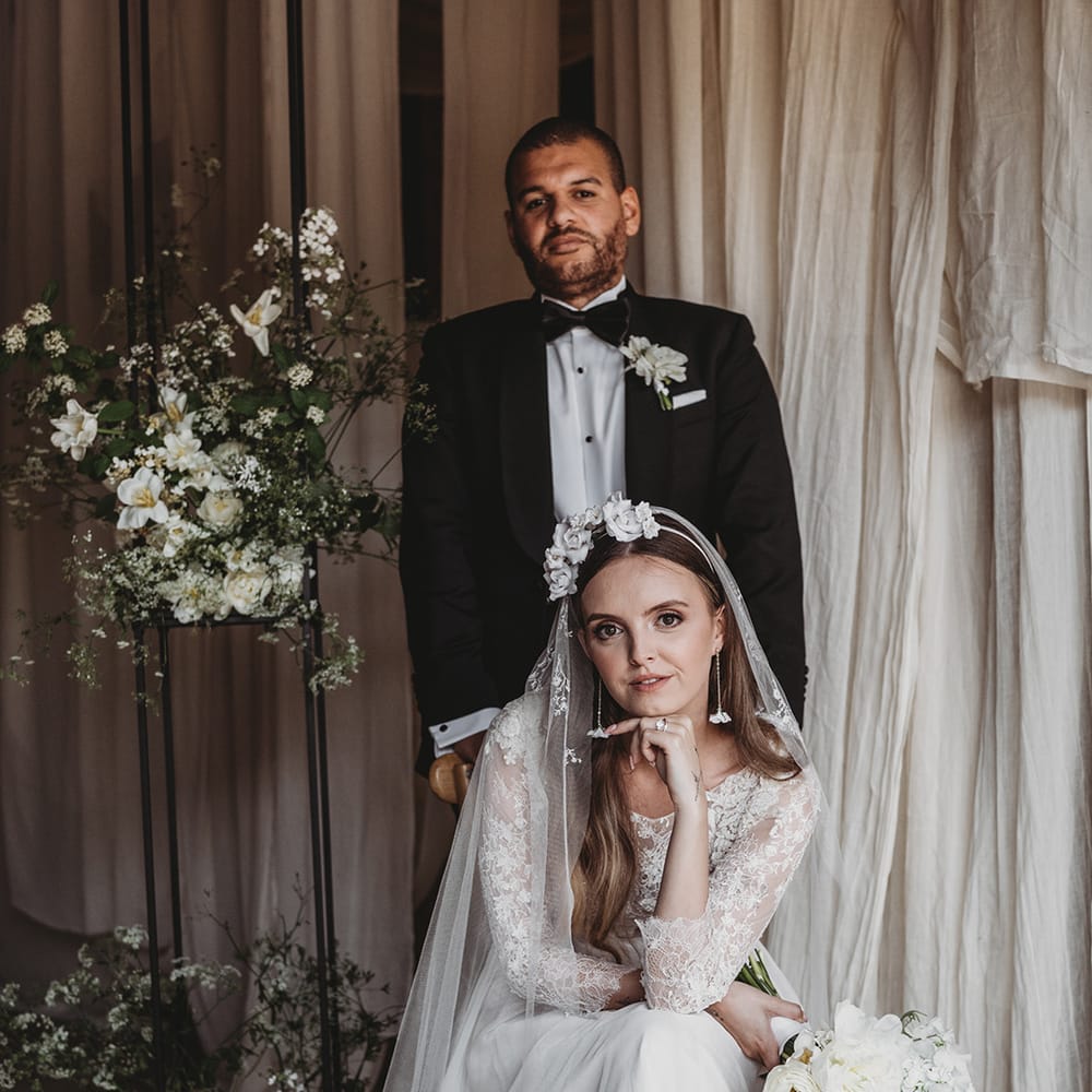 Bride in lace wedding dress, embroidered veil, floral headband and earrings with groom in black tie 