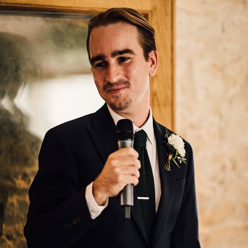 Groom stands with a microphone to deliver a speech in a navy blue suit and white rose buttonhole 