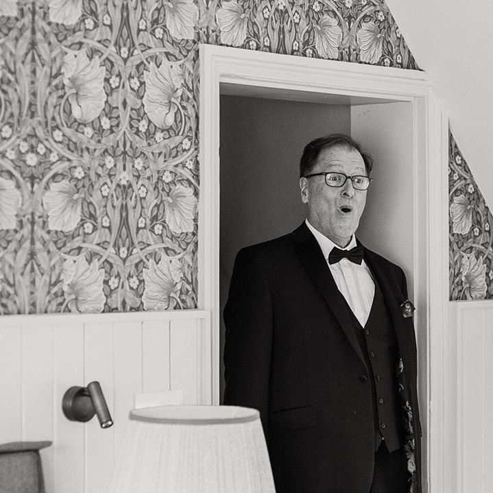 Father of the bride in black tie has a shocked facial expression as he sees the bride for the first time 