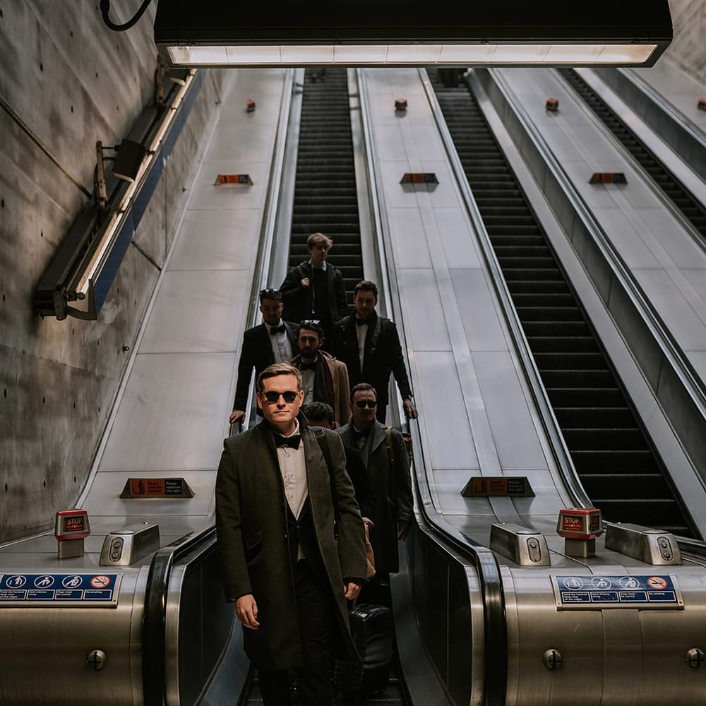 Grooms party in matching black tuxedos and sunglasses going down the escalator in the London Tube