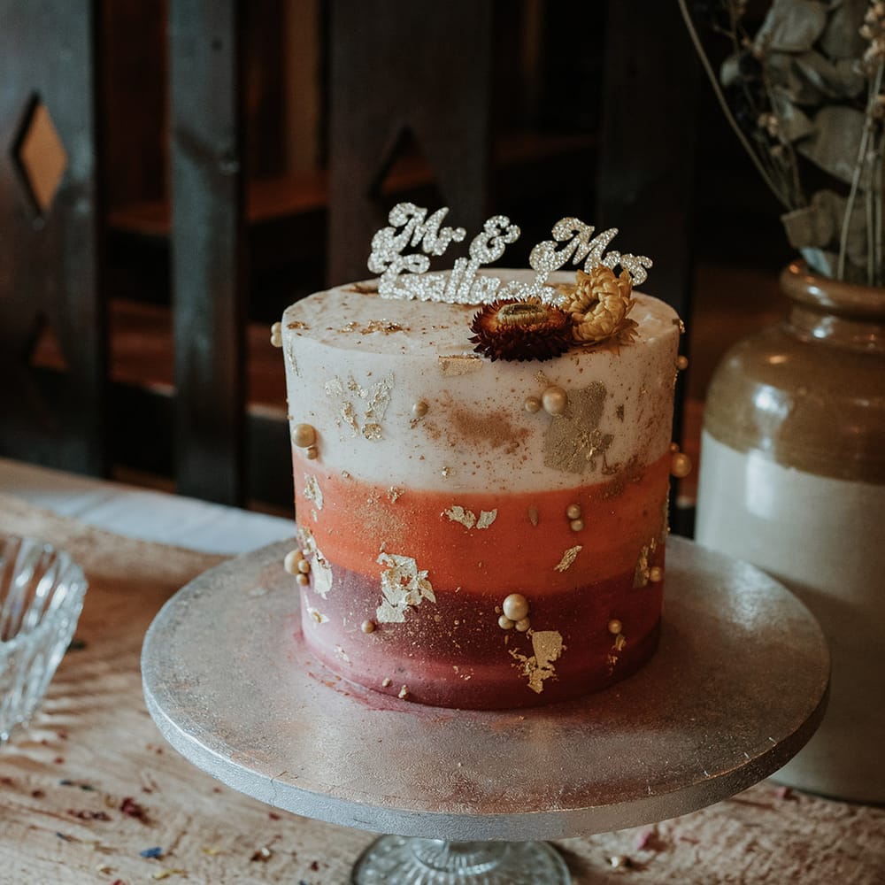 Single tier buttercream wedding cake in white, orange and pink on stone cake stand with personalised glittery wedding cake topper
