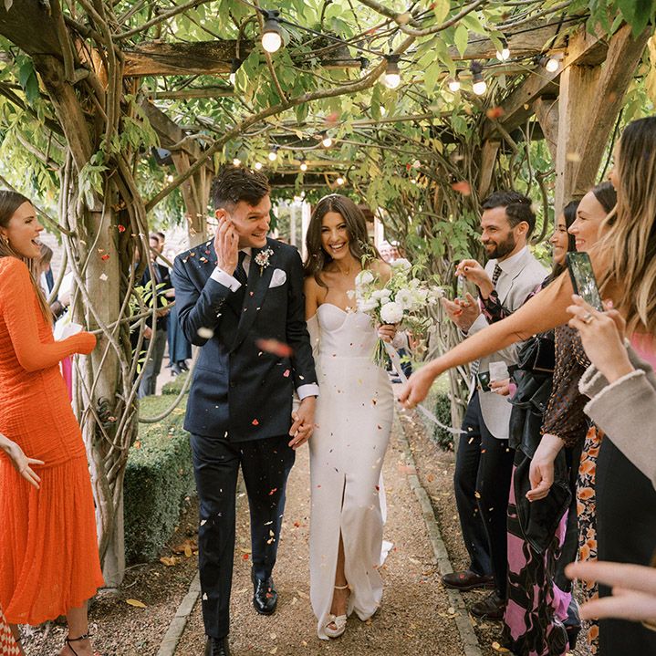Groom in a dark suit walking with the bride in a fitted wedding dress with split leg to their confetti exit 