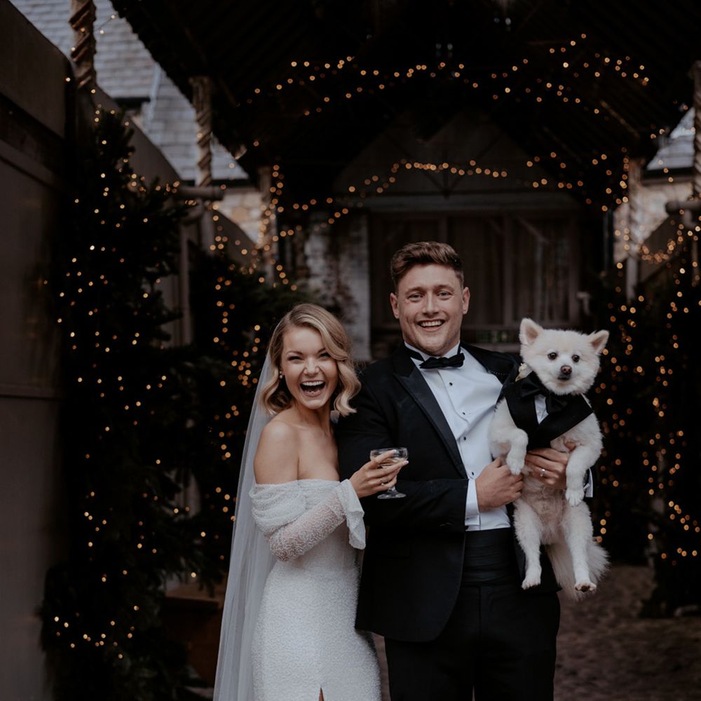 The bride and groom smiling with their pet dog wearing a tuxedo dog wedding outfit 