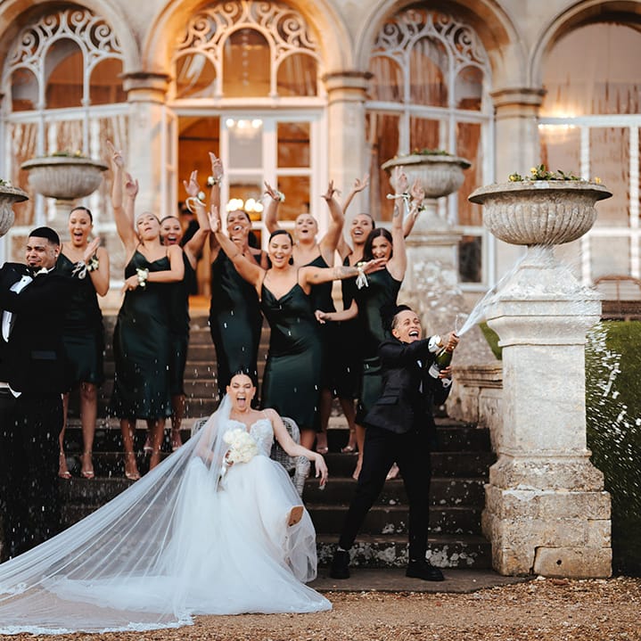 Champagne pop wedding photoshoot with bridesmaids in satin black strappy bridesmaid dresses and classic black tuxedos, bride in black snakeskin print tuxedo with black bowtie and white rose boutonniere and bride in lace sleeveless v neck Enzoani wedding dress with tulle finish and church length veil at Grittleton House 
