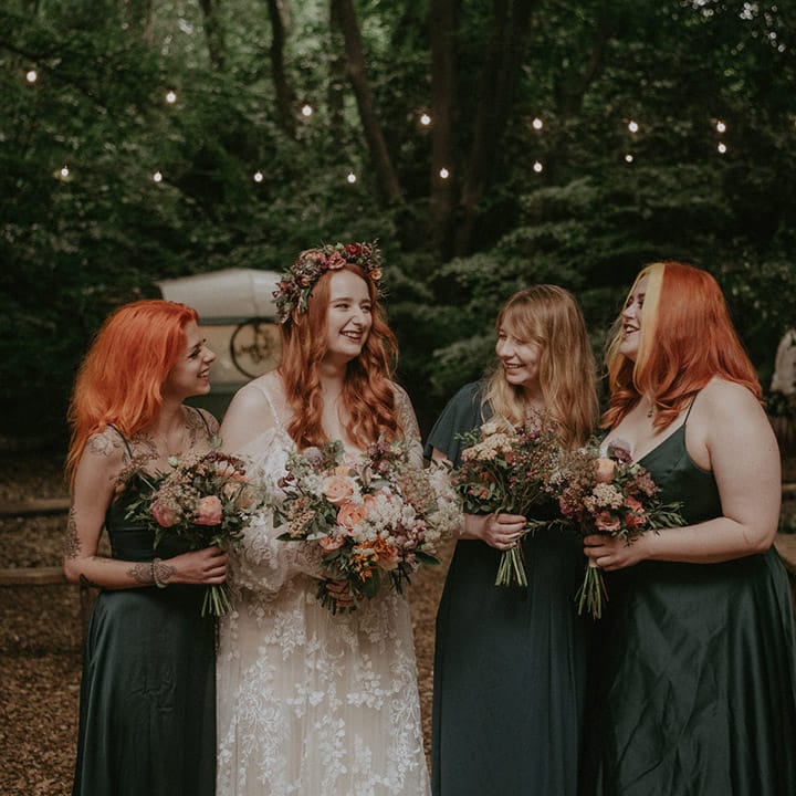 Bride in off the shoulder boho lace wedding dress and pink flower crown with the bridesmaids in green satin bridesmaid dresses 