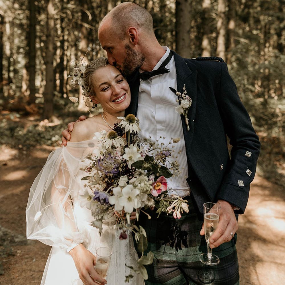Groom in bespoke checkered trousers and pleated shirt kissing bride on the forehead holding a wildflower wedding bouquet 
