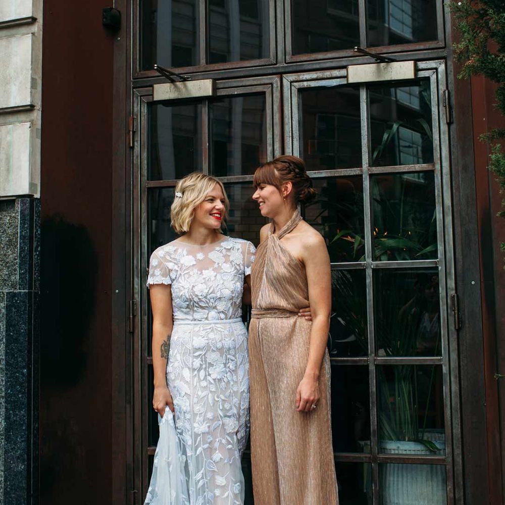 Bride in lace short sleeve wedding dress standing with bridesmaid in gold sparkly halterneck jumpsuit - hen party ideas