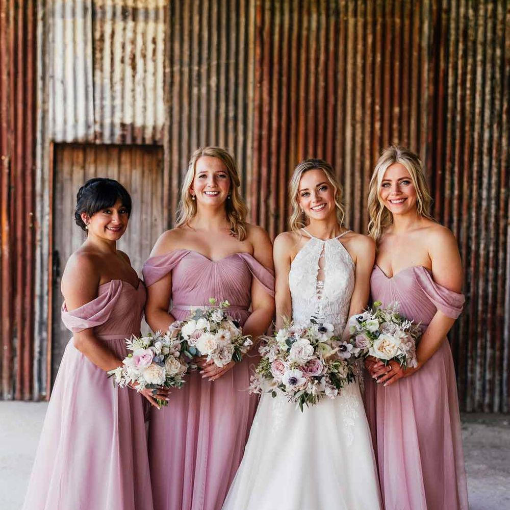 Bride wearing cut out sleeveless lace wedding dress standing with bridesmaids in off the shoulder dusky pink bridesmaid dresses 