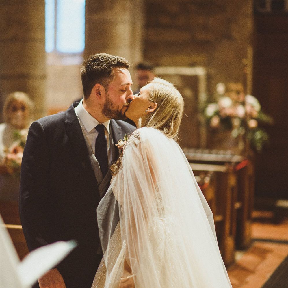 Bride with side swept hair and wedding cape kissing her husband in the church 