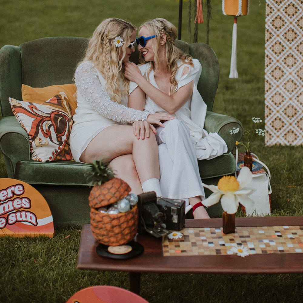 Two brides in sunglasses embracing on a green velvet loveseat at sixties wedding 