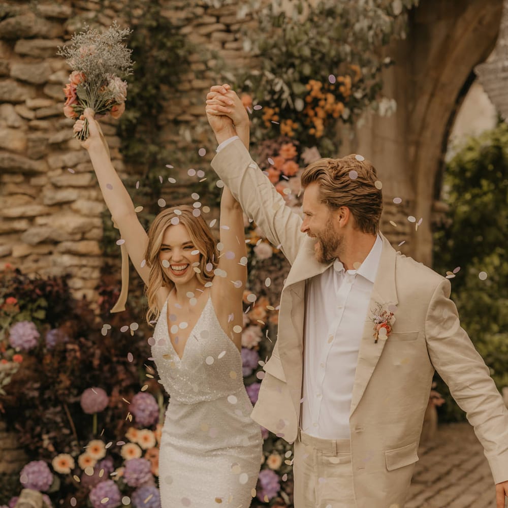 Groom in a beige suit and white shirt holding hands with his bride in a sparkly wedding dress as they wavy hands and celebrate as confetti is thrown over them