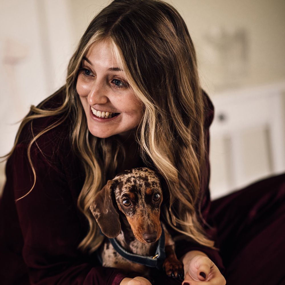 Bride in burgundy pyjamas with curled hair hugs miniature daschund before church wedding ceremony in Cornwall