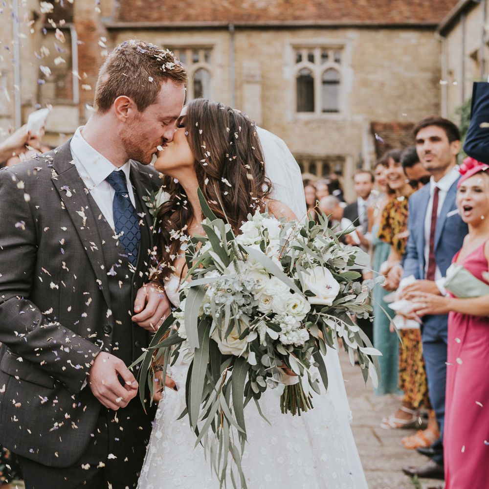 Bride in Pronovias wedding dress and veil holding white and green bridal bouquet kisses groom in grey three piece suit, blue tie and rose buttonhole as guests throw confetti at Notley Abbey wedding