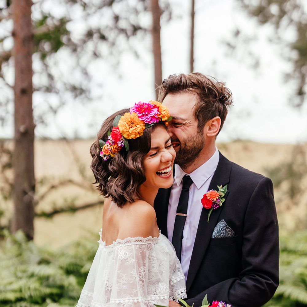 Bride in off the shoulder Grace Loves Lace wedding dress and flower crown holding multicoloured wedding bouquet laughs as she hugs groom in black suit and colourful floral buttonhole in woodland