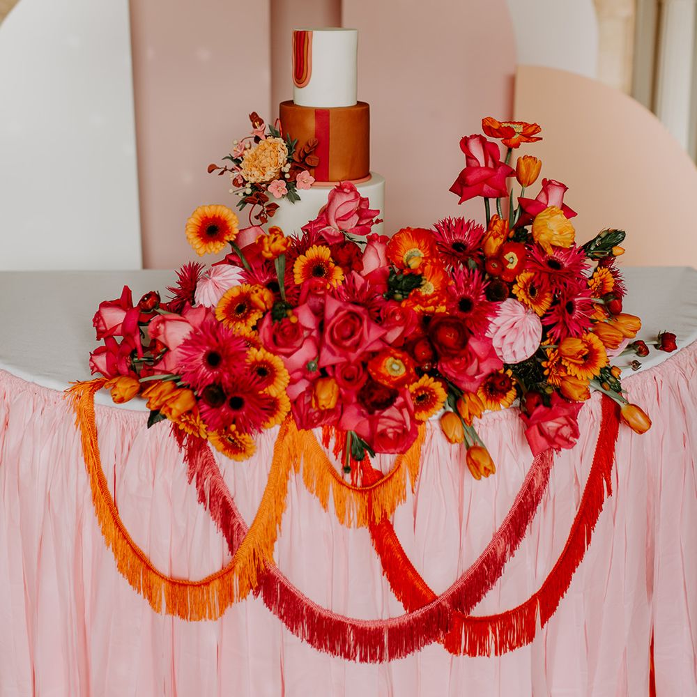 Wedding cake table with pink tablecloth and geometric backdrop and vibrant red floral arrangement and tassels 
