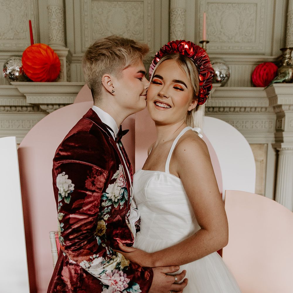 Bride in a burgundy velvet jacket with floral design kissing her bride in a tulle skirt wedding dress and red applique headband 