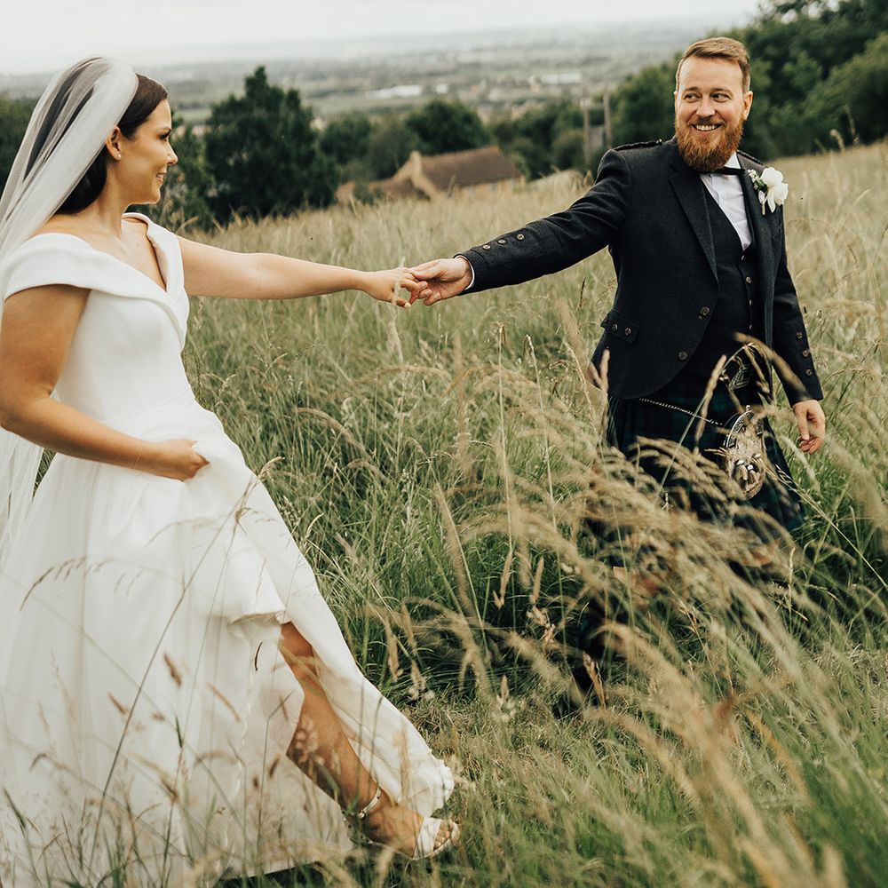 Groom in a tartan kilt holding hands with his bride in a Suzanne Neville princess wedding dress in field 
