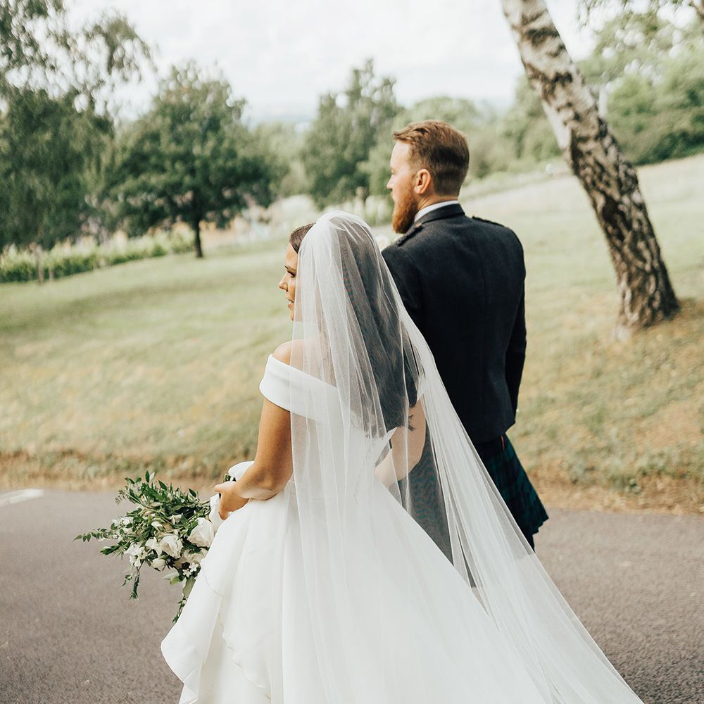 Bride in a Suzanne Neville off the shoulder wedding dress and long veil walking hand in hand with her groom in a tartan kilt 