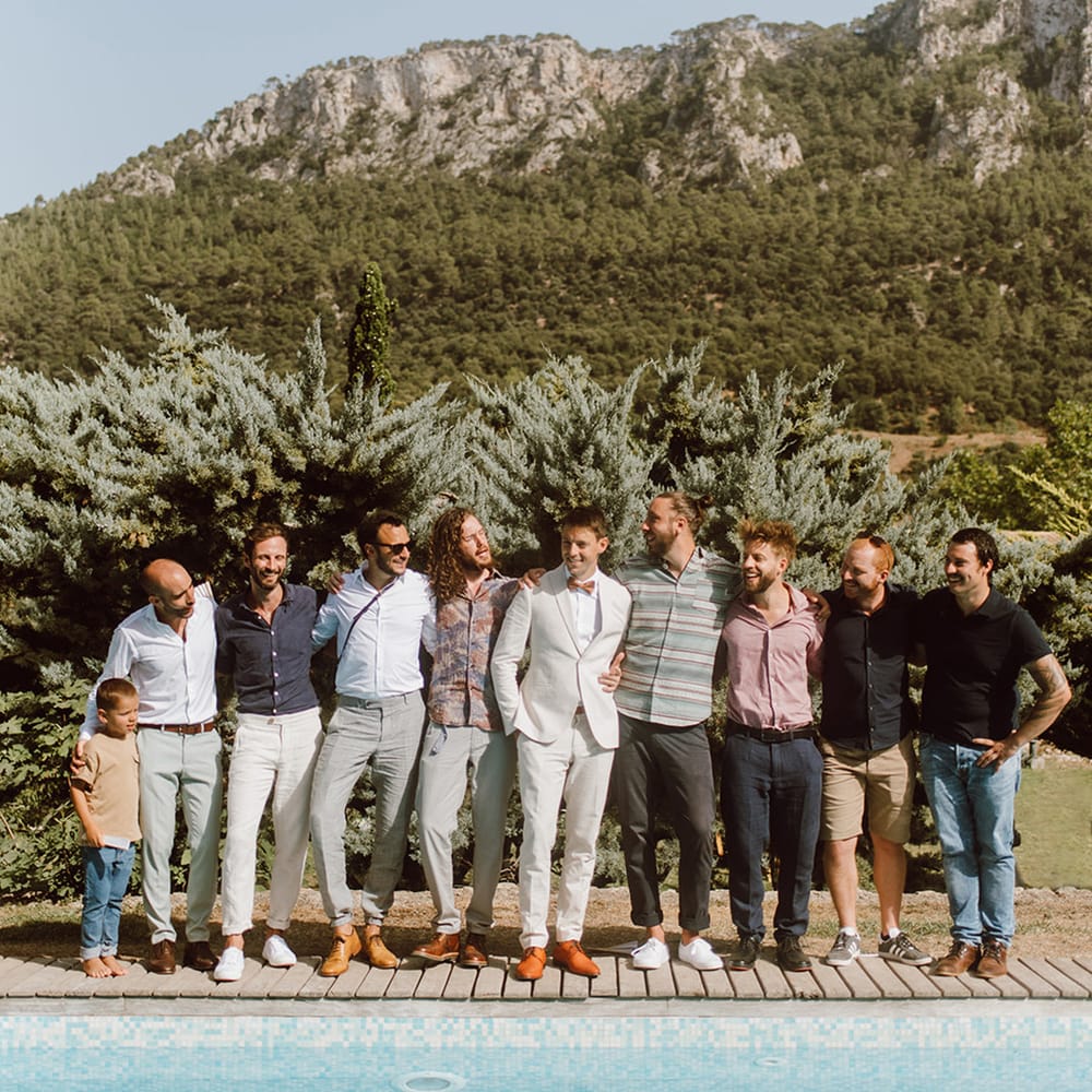 groom in beige suit and pink bow tie smiles with groomsmen in front of a pool in Mallorca